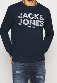 Marinblå sweatshirt med ribbad mudd och nederkant, med fet vit text "JACK & JONES EST. 1990" över bröstet. Blandning av bomull.