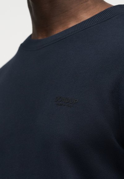 Κοντινό πλάνο ενός ατόμου που φοράει πουλόβερ σκούρου ναυτικού μπλε με λαιμόκοψη, με την εγχάραξη «DONDUP Made in Italy» στο στήθος.