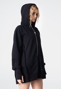 Czarna bluza z kapturem zapinana na zamek, wykonana z miękkiego materiału, z dużym kapturem, przednimi kieszeniami i mankietami. Szczegóły tekstowe na podszewce kaptura.