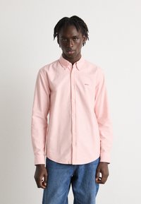 Camicia button-down rosa chiaro con colletto, caratterizzata da un piccolo logo "BOSS". Il tessuto sembra liscio e leggermente strutturato. Indossata con jeans blu.