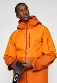 Mies, jolla on päällä oranssi huppari, säätää mustia hiihtolasit, joissa on valkoinen Oakley-logo hihnassa.