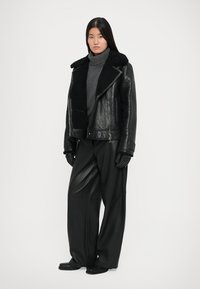 Črna usnjena motoristična jakna z volnenim ovratnikom, oblečena čez sivo turtleneck majico, skupaj s širokimi črnimi hlačami in črnimi rokavicami.