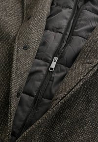 Brun fuskpälsrock med svart quiltad foder med dragkedja, två knappar framtill och en slät textur med kontrasterande material.