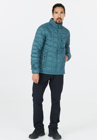 Teal quiltet jakke med høj krave, fuld lynlås og sidelommer. Båret med sorte bukser og mørke sko. Ensartet tekstur, praktisk design.