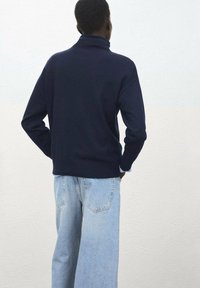 Granatowy golf z miękkiej tkaniny, zestawiony z jasnoniebieskimi dżinsami o szerokich nogawkach; widok od tyłu na tle neutralnej ściany.