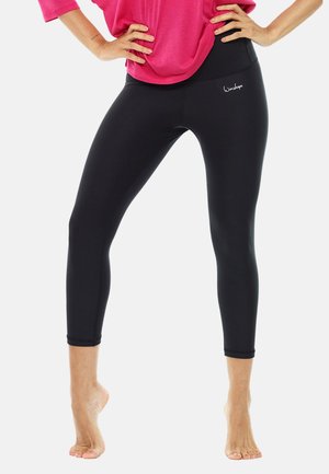 Kvinna i svarta åtsittande korta leggings och en rosa topp, stående barfota med händerna på höfterna mot en enfärgad bakgrund.