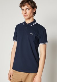 Námornícky polo tričko vyrobené z bavlny s trojbuttonovou légou, bielym pruhovaným golierom a manžetami, a vyšitým logom "BOSS" na hrudi.
