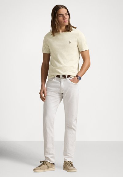 Světle žluté tričko s krátkým rukávem a vyšívaným logem, doplněné bílými džínami a béžovými teniskami. Model stojí na neutrálním pozadí.