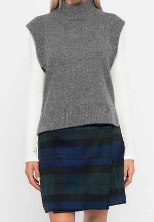 Kobieta w szarym bezrękawniku z dzianiny z golfem na białej bluzce z długimi rękawami, w połączeniu z zielono-niebieską spódnicą w kratę.