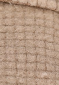 Tessuto beige, strutturato, con un motivo a griglia e una superficie morbida e pelosa. Il materiale sembra essere infeltrito o tessuto, mostrando una sottile morbidezza.