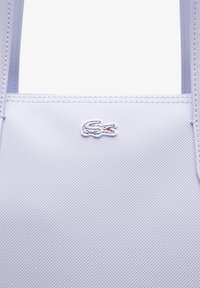 Bolso tote sintético de color púrpura claro con superficie texturizada, que presenta un prominente logo de cocodrilo bordado en blanco y asas resistentes.