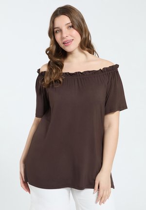 Femme aux cheveux ondulés portant un haut marron épaules dénudées et un pantalon blanc, souriant légèrement sur un fond clair uni.