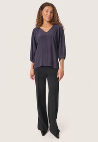 Blusa fluida di un viola scuro con scollatura a V e maniche arricciate, abbinata a pantaloni neri a vita alta e décolleté neri appuntiti.