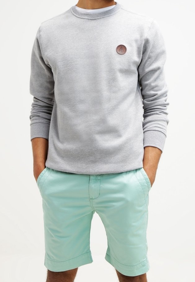 Mann in einem hellgrauen Langarm-Sweatshirt mit rundem Emblem und hellblauen, knielangen Shorts, die Hände in den Taschen.