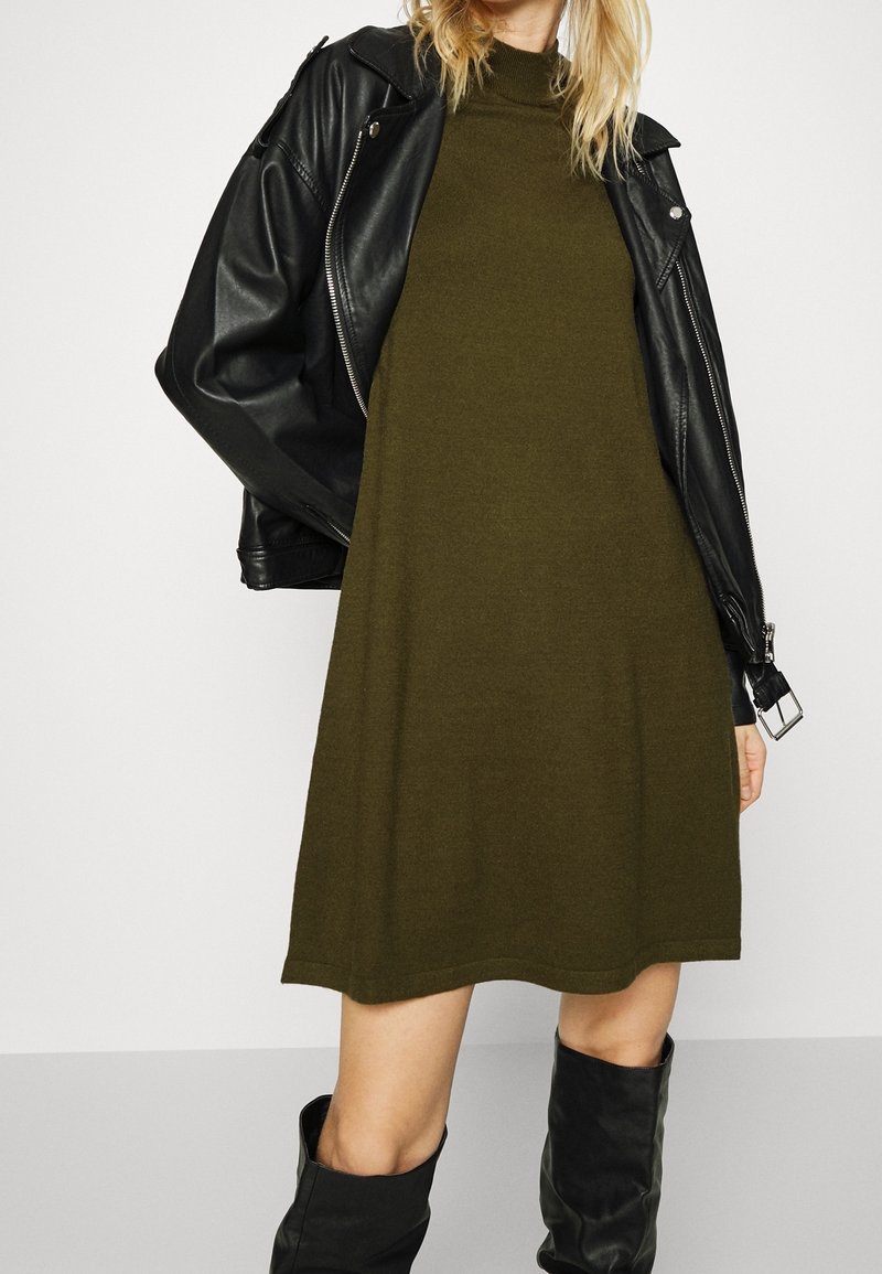 Olivgrön stickad klänning med hög halsringning, tillsammans med en svart lädermotorcykeljacka. Outfiten inkluderar knähöga svarta stövlar.