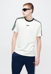 Noor mees, kes kannab musti päikeseprille, valget Adidas t-särki triibuliste varrukatega ja tumesiniseid Adidas dressipükse, seisab käed taskutes.