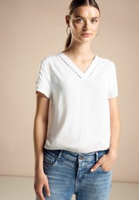 Blusa bianca a maniche corte con scollatura a V in tessuto liscio, caratterizzata da cuciture decorative lungo il collo, abbinata a jeans in denim blu.