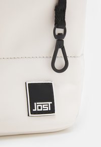 Clip noir attaché à une sangle noire sur un sac texturé blanc avec une étiquette carrée noire comportant un texte blanc "Jost".