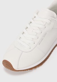 White leather sneaker with a rounded toe, featuring flat white laces, a textured rubber sole, and subtle gray branding on the side.