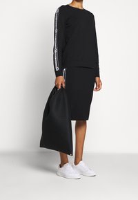 Czarny długi rękaw bluzę z logo w paski, zestawioną z dopasowaną czarną spódnicą. Białe sneakersy i fakturowana czarna torebka dopełniają stylizację.