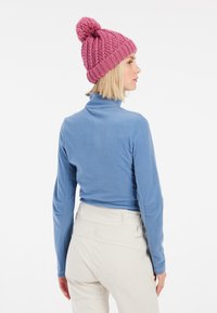 Maglione in fleece blu con colletto alto, abbinato a un berretto lavorato a maglia rosa con pom-pom, e pantaloni di colore chiaro.