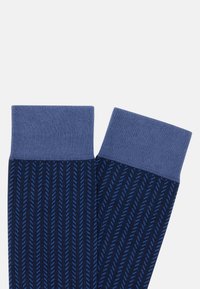 Tumšzilgu zeķu pāris ar gaiši zilgu ķežonu rakstu un vienkāršām gaiši zilgām aprocēm, izklāts uz baltas fona.