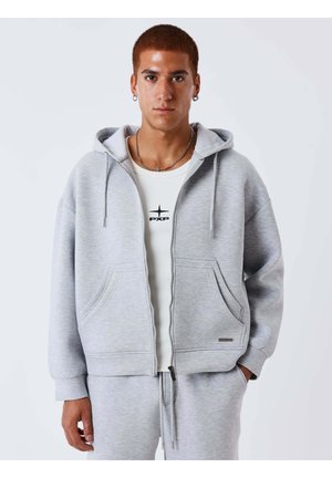 Jeune homme aux cheveux roux courts portant des boucles d'oreilles argentées en forme d'anneau, un sweat à capuche zippé gris clair, un pantalon de survêtement assorti et un t-shirt blanc avec un logo.