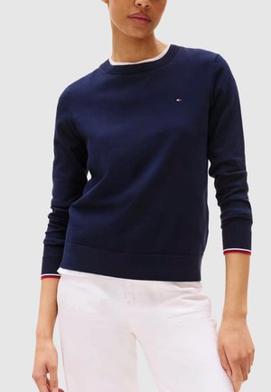 Persona che indossa un maglione a maniche lunghe blu navy con dettagli bianchi e rossi ai polsini e al collo, abbinato a pantaloni bianchi, su uno sfondo uniforme.