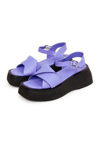 Sandali sintetici viola con cinturini incrociati, fibbia alla caviglia regolabile e suola nera rialzata. Presentano una soletta piatta e dettagli di cucitura.