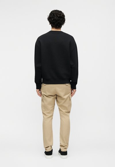 Άτομο που στέκεται με την πλάτη στην κάμερα, φοράει μαύρο μακρυμάνικο φούτερ, μπεζ παντελόνι cargo και μαύρα αθλητικά παπούτσια με λευκές σόλες.