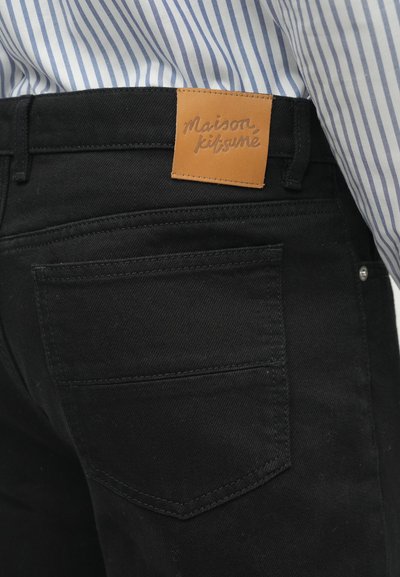 Jeans negre din denim, cu un eticheta de piele maro cu text embossat. Buzunarul din spate are un design clasic și detalii vizibile de cusătură.