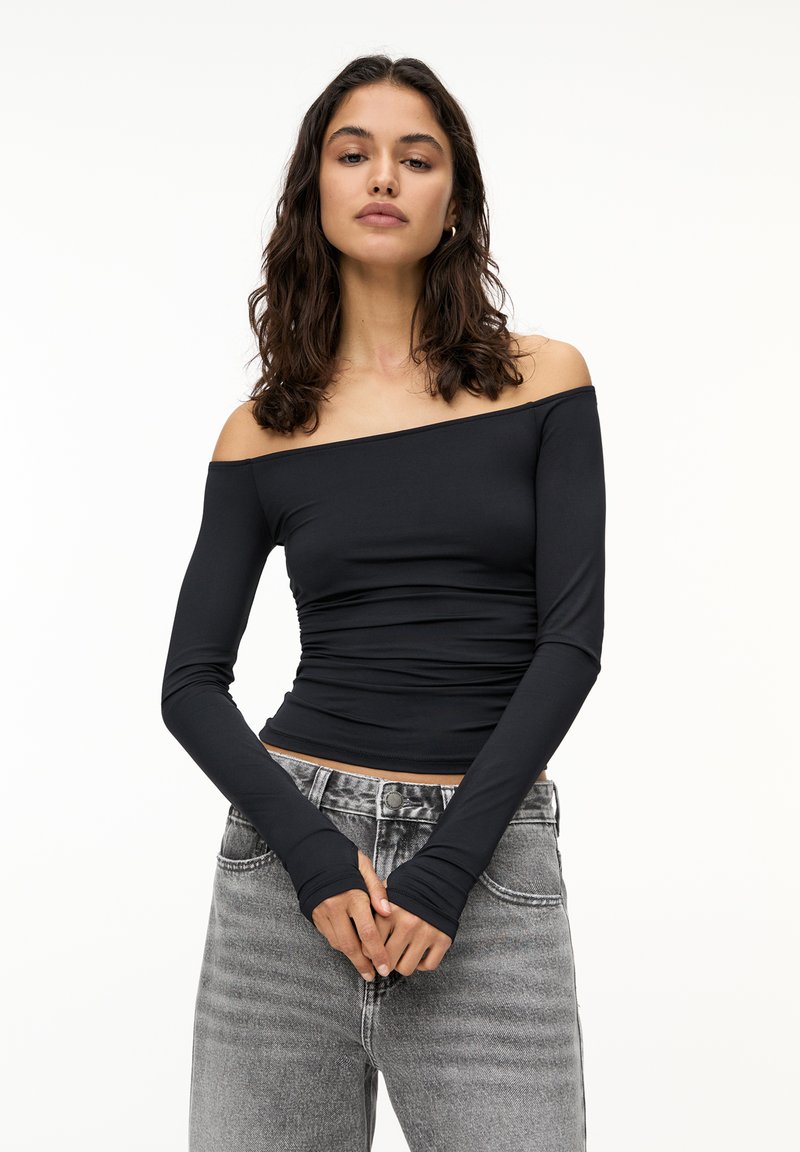 Schwarzes, schulterfreies Oberteil mit langen Ärmeln und Raffung; getragen mit hellgrauen, hoch geschnittenen Jeans in ausgewaschenem Design.