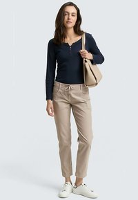 Granatowy długi rękaw koszulki henley, beżowe spodnie z trokami i białe trampki. Model trzyma beżową torbę na ramię; neutralne tło.