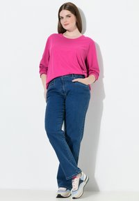Różowa bluzka z długim rękawem, niebieskie dżinsy z kieszeniami oraz wielokolorowe buty sportowe z masywną podeszwą, na białym tle.