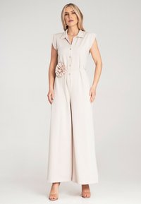 Beżowy kombinezon z krótkimi rękawami, kołnierzykiem, guzikami z przodu i kwiatowym akcentem w talii. Szerokie nogawki, gładka tekstura materiału.