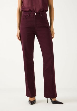 Vrouw draagt donker bordeauxrode jeans met hoge taille en rechte pijpen, gecombineerd met zwarte spitse hoge hakken en een bijpassende bordeauxrode top.