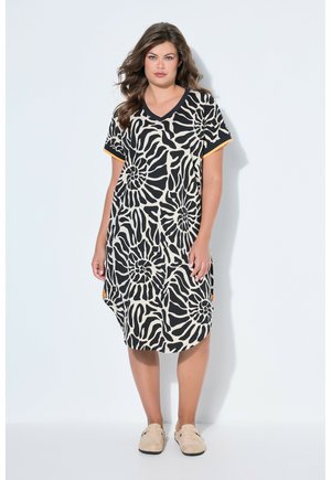 Femme portant une robe noire et blanche à manches courtes avec des motifs spirales zébrés, debout devant un fond clair uni, chaussures beiges.