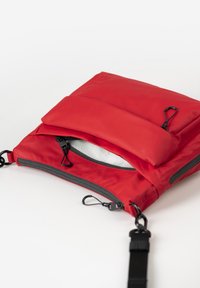 Rote Stofftasche mit zwei Reißverschlüssen, einer Vordertasche und einem schwarzen Trageriemen. Besitzt eine glatte Textur und kontrastierende graue Reißverschlussdetails.