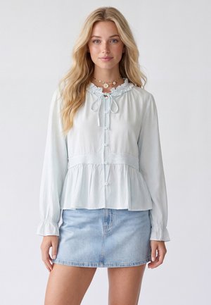 Ung kvinde med langt blondt hår iført en hvid flæset bluse med knapper og en lyseblå denim nederdel mod en ensfarvet baggrund.