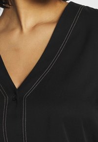 Top nero a scollo a V realizzato in tessuto liscio con dettagli di cucitura rosa sottile e un bottone sul collo, caratterizzato da una vestibilità comoda.