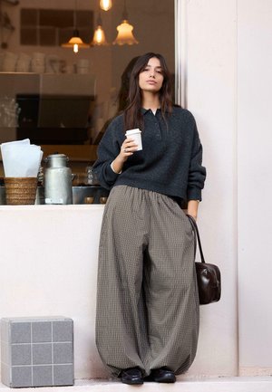 Jeune femme appuyée contre un mur à l'extérieur d'un café, tenant une tasse de café dans une main et un sac à main marron foncé dans l'autre.