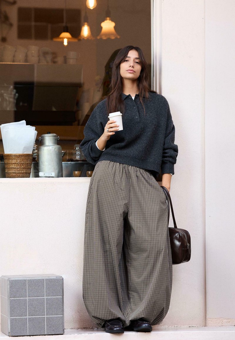 Jeune femme appuyée contre un mur à l'extérieur d'un café, tenant une tasse de café dans une main et un sac à main marron foncé dans l'autre.