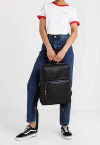 Svart faux läder ryggsäck med en framficka med dragkedja, justerbara remmar och en minimalistisk design. Bärs med en vit t-shirt och blå jeans.