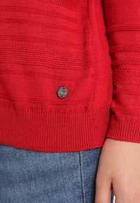 Rode gebreide trui met horizontale textuurpatronen en geribbelde manchetten. Voorzien van een cirkelvormig grijs label met het woord "Esprit." Gecombineerd met denim.