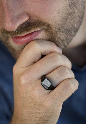 Schwarzer Metallring mit einem rechteckigen Stein, der in der Mitte eingelegt ist und eine grau-weiße gemusterte Textur aufweist. Hand ruht nahe am Kinn.
