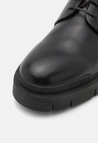 Černá kožená bota s hladkým svrškem, kulatou špičkou a silnou černou gumovou podrážkou se strukturovaným akcentem a detailem značky.