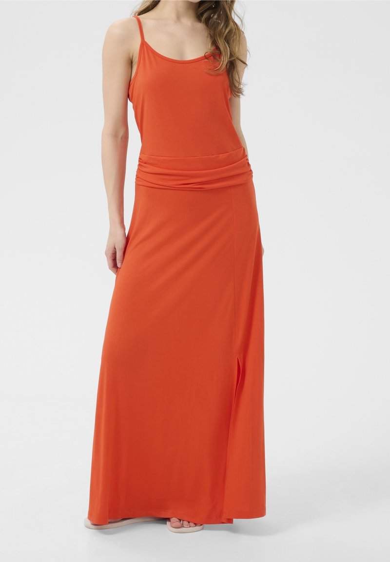 Femme portant une robe longue orange sans manches avec de fines bretelles et une taille froncée, debout devant un fond clair uni.
