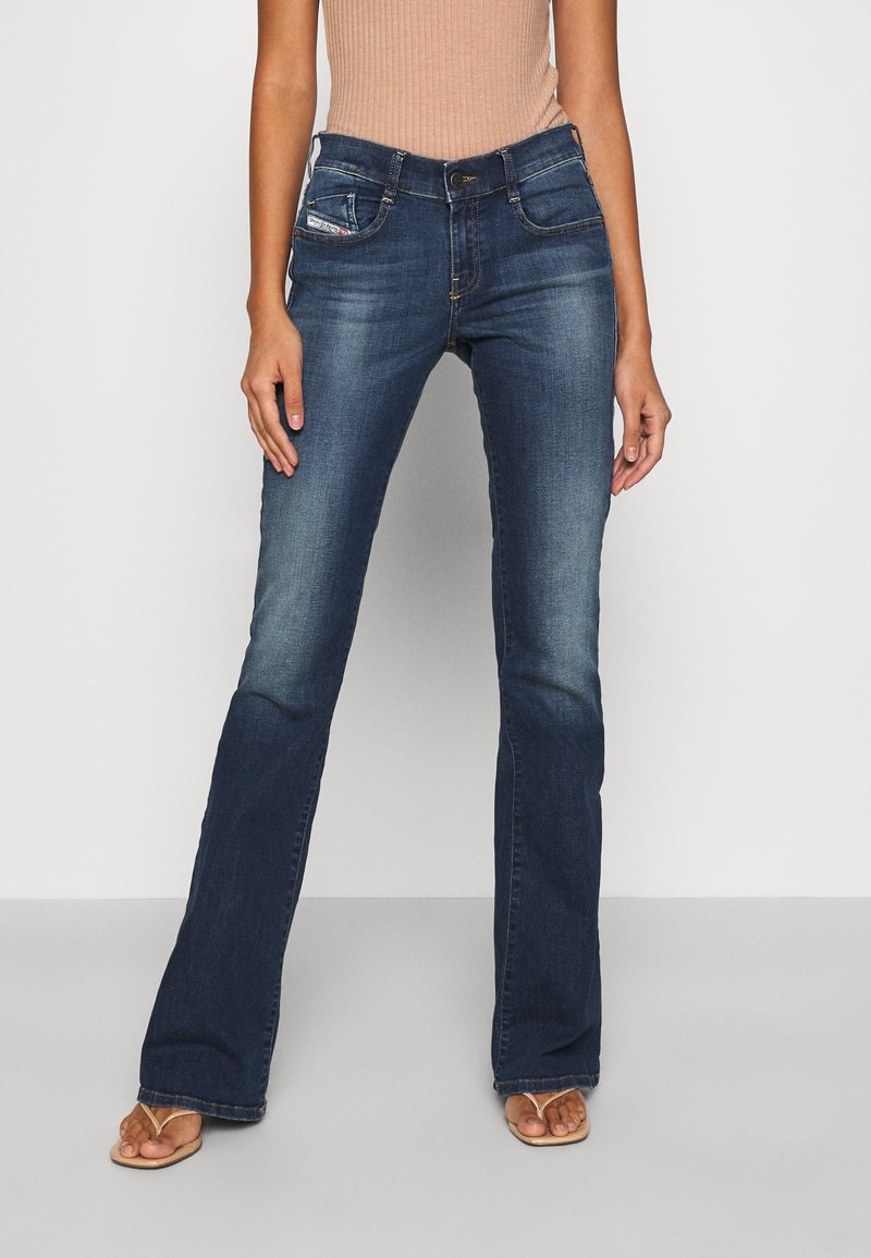 Femme portant un jean bootcut bleu foncé avec décoloration aux cuisses et aux genoux, associé à un top côtelé sans manches beige et des sandales beiges.
