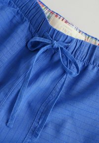 Pantalon en lin bleu avec taille élastique et cordon noué, montrant une doublure en tissu à carreaux et une étiquette indiquant la teneur en lin.