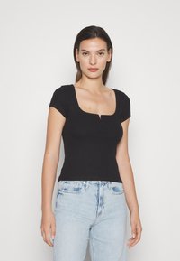 Top aderente nero a maniche corte con scollo quadrato e un leggero dettaglio con apertura al centro, realizzato in tessuto a coste. Abbinato a jeans azzurri chiari.